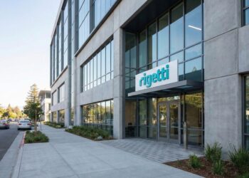 Fotografía exterior a nivel de calle de un edificio moderno de oficinas de hormigón y vidrio. Un letrero prominente con el logo azul y el texto "Rigetti" está ubicado sobre la entrada principal. La escena es durante el día, con una acera limpia y una calle tranquila con algunos vehículos, bajo un cielo despejado.
