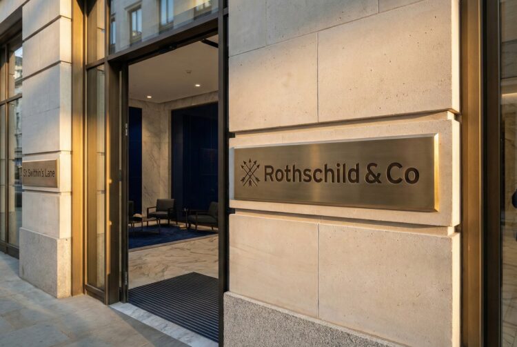 Fotografía exterior de la entrada principal de la sede corporativa de la banca de inversión Rothschild & Co en New Court, St Swithin's Lane, Londres. Una placa de bronce con el nombre de la firma y el logo de las cinco flechas destaca sobre la fachada de piedra caliza iluminada por el sol del atardecer, con un vistazo al moderno vestíbulo interior.