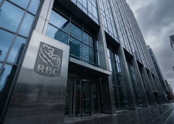 Fotografía en ángulo contrapicado de la entrada principal de la torre de oficinas de RBC. Un gran letrero de metal cepillado con el logo y las siglas "RBC" destaca junto a las puertas giratorias de vidrio. El moderno rascacielos de cristal y acero se alza bajo un cielo denso y gris, con el pavimento mojado reflejando la luz fría en un distrito financiero.