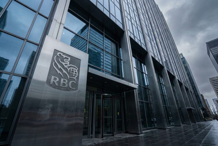 Fotografía en ángulo contrapicado de la entrada principal de la torre de oficinas de RBC. Un gran letrero de metal cepillado con el logo y las siglas "RBC" destaca junto a las puertas giratorias de vidrio. El moderno rascacielos de cristal y acero se alza bajo un cielo denso y gris, con el pavimento mojado reflejando la luz fría en un distrito financiero.