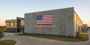 Fotografía exterior del edificio de la empresa USA Rare Earth. La imagen muestra una instalación industrial moderna con una gran bandera estadounidense y el nombre "USA Rare Earth" en letras metálicas en la fachada principal. El sol del atardecer ilumina el edificio y el entorno, que incluye un camino de entrada pavimentado y áreas verdes.