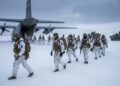 Soldados de EE. UU. y de la OTAN, vestidos con camuflaje de invierno completo, mochilas y rifles, caminan en fila alejándose de la rampa trasera de un avión de transporte militar C-130 Hercules en una llanura cubierta de nieve bajo un cielo nublado. Se ven vehículos militares en la distancia. Los parches en sus brazos indican diferentes nacionalidades, incluidas las de EE. UU. y Noruega.