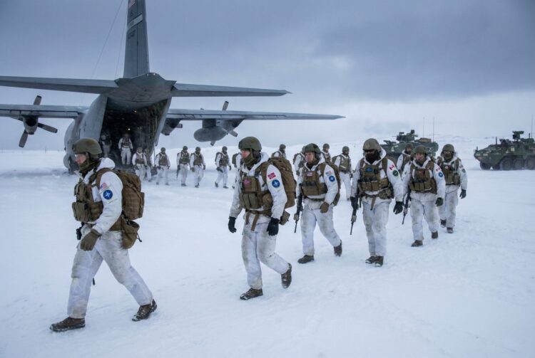 Soldados de EE. UU. y de la OTAN, vestidos con camuflaje de invierno completo, mochilas y rifles, caminan en fila alejándose de la rampa trasera de un avión de transporte militar C-130 Hercules en una llanura cubierta de nieve bajo un cielo nublado. Se ven vehículos militares en la distancia. Los parches en sus brazos indican diferentes nacionalidades, incluidas las de EE. UU. y Noruega.