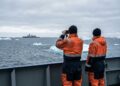 Una fotografía documental muestra a dos hombres de espaldas con trajes de trabajo naranja y azul marino en la cubierta de un barco. Uno usa prismáticos para observar un buque militar gris distante en un mar frío y gris salpicado de icebergs bajo un cielo nublado.