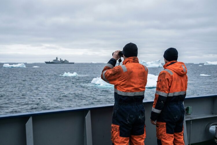 Una fotografía documental muestra a dos hombres de espaldas con trajes de trabajo naranja y azul marino en la cubierta de un barco. Uno usa prismáticos para observar un buque militar gris distante en un mar frío y gris salpicado de icebergs bajo un cielo nublado.