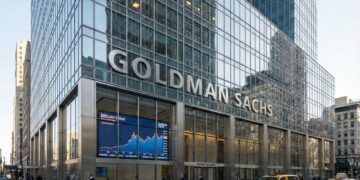 Una fotografía del exterior del rascacielos sede de Goldman Sachs en 200 West Street en la ciudad de Nueva York. El moderno edificio de fachada de vidrio y acero presenta grandes letras metálicas que dicen "GOLDMAN SACHS". A nivel de calle, una gran pantalla digital dentro de una ventana muestra un gráfico titulado "Bitcoin Price". Peatones caminan por la acera y un taxi amarillo circula por la calle. La fachada del edificio refleja otras estructuras de la ciudad bajo la luz del día.