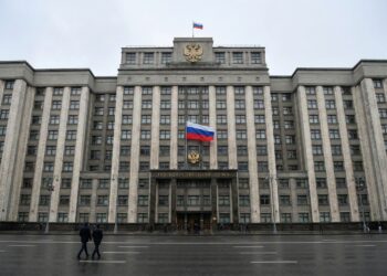 Una fotografía de paisaje de ángulo amplio muestra el edificio de la Duma Estatal en Moscú en un día nublado, con una gran bandera rusa y el escudo de armas sobre la entrada principal, con asfalto mojado y algunos peatones en primer plano.