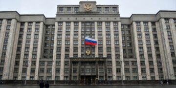 Una fotografía de paisaje de ángulo amplio muestra el edificio de la Duma Estatal en Moscú en un día nublado, con una gran bandera rusa y el escudo de armas sobre la entrada principal, con asfalto mojado y algunos peatones en primer plano.