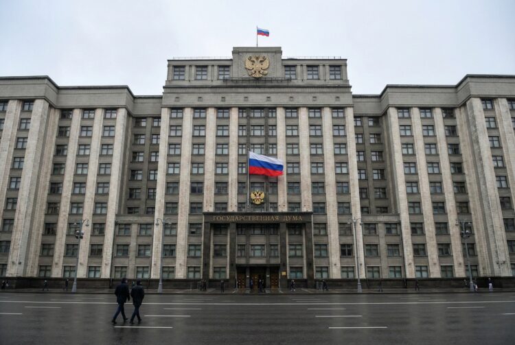 Una fotografía de paisaje de ángulo amplio muestra el edificio de la Duma Estatal en Moscú en un día nublado, con una gran bandera rusa y el escudo de armas sobre la entrada principal, con asfalto mojado y algunos peatones en primer plano.