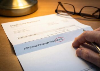 Fotografía de primer plano sobre un escritorio de madera que muestra un estado de cuenta de tarjeta de crédito impreso. Una mano sostiene un bolígrafo y señala la línea de texto que dice "APR (Annual Percentage Rate):", donde el valor numérico "10.00%" está claramente marcado dentro de un círculo rojo manuscrito. Unas gafas de lectura y la base de una lámpara están desenfocadas en el fondo bajo una iluminación cálida.
