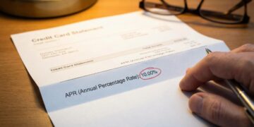 Fotografía de primer plano sobre un escritorio de madera que muestra un estado de cuenta de tarjeta de crédito impreso. Una mano sostiene un bolígrafo y señala la línea de texto que dice "APR (Annual Percentage Rate):", donde el valor numérico "10.00%" está claramente marcado dentro de un círculo rojo manuscrito. Unas gafas de lectura y la base de una lámpara están desenfocadas en el fondo bajo una iluminación cálida.