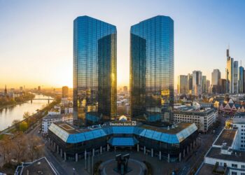 Fotografía panorámica de las Torres Gemelas del Deutsche Bank en Frankfurt, Alemania, al amanecer. Los dos rascacielos de vidrio azul reflejan la luz dorada del sol naciente, que brilla intensamente entre ellos. El logotipo de "Deutsche Bank" es visible en la base del edificio. El río Meno y el horizonte de la ciudad de Frankfurt, con otros edificios y puentes, se extienden en el fondo bajo un cielo despejado que pasa de naranja a azul.
