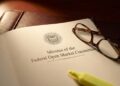 Fotografía de primer plano del documento oficial impreso con el título "Minutes of the Federal Open Market Committee" y el sello del Sistema de la Reserva Federal. Unas gafas de lectura con montura de carey y un rotulador amarillo fluorescente descansan sobre la página, que se encuentra sobre un portafolios de cuero oscuro bajo una luz focalizada.