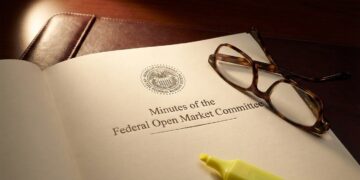 Fotografía de primer plano del documento oficial impreso con el título "Minutes of the Federal Open Market Committee" y el sello del Sistema de la Reserva Federal. Unas gafas de lectura con montura de carey y un rotulador amarillo fluorescente descansan sobre la página, que se encuentra sobre un portafolios de cuero oscuro bajo una luz focalizada.