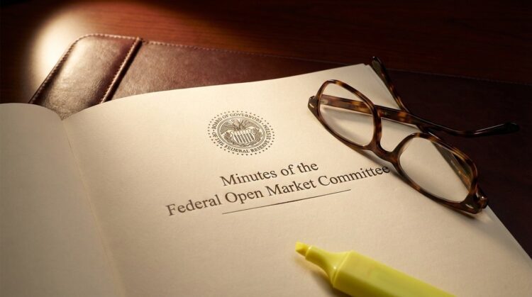 Fotografía de primer plano del documento oficial impreso con el título "Minutes of the Federal Open Market Committee" y el sello del Sistema de la Reserva Federal. Unas gafas de lectura con montura de carey y un rotulador amarillo fluorescente descansan sobre la página, que se encuentra sobre un portafolios de cuero oscuro bajo una luz focalizada.