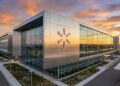Fotografía arquitectónica panorámica del moderno edificio de la sede corporativa de Walmart. La fachada, compuesta por paneles de metal cepillado y grandes muros cortina de vidrio, refleja los tonos naranjas y dorados de la puesta de sol. El gran logotipo de la "chispa" (spark) de Walmart en relieve metálico es prominente en la esquina principal del edificio. El entorno incluye pasarelas pavimentadas, jardines cuidados y estructuras de paneles solares en el estacionamiento al fondo.