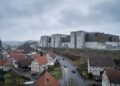 Fotografía panorámica de un pequeño pueblo alemán con casas de entramado de madera y una iglesia con campanario bajo un cielo nublado. Una carretera sinuosa con algunos coches atraviesa el pueblo. En el fondo, sobre una colina, se alza un enorme edificio brutalista de hormigón gris y paneles oxidados, dominando el paisaje rural.