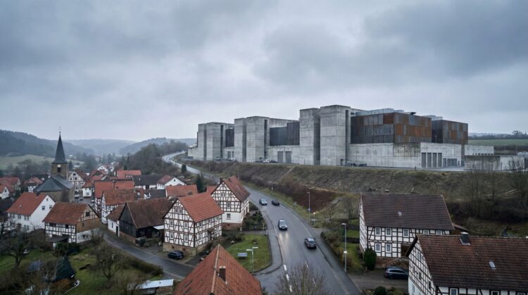 Fotografía panorámica de un pequeño pueblo alemán con casas de entramado de madera y una iglesia con campanario bajo un cielo nublado. Una carretera sinuosa con algunos coches atraviesa el pueblo. En el fondo, sobre una colina, se alza un enorme edificio brutalista de hormigón gris y paneles oxidados, dominando el paisaje rural.