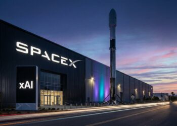 Fotografía de larga exposición al atardecer de la fachada de la sede corporativa de SpaceX en Hawthorne, California. El edificio industrial presenta el logotipo de 'SpaceX' iluminado en blanco, mientras que un tótem digital adyacente muestra prominentemente el logotipo de la empresa de inteligencia artificial 'xAI'. Un cohete propulsor Falcon 9 está expuesto verticalmente frente a las instalaciones, con estelas de luz de tráfico de vehículos en primer plano.