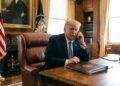 Una fotografía editorial cándida muestra a Donald Trump sentado en un sillón de cuero marrón en una oficina clásica con paneles de madera. Está hablando por un teléfono de cable con una expresión seria. Sobre el escritorio de madera hay una carpeta de cuero con el sello presidencial. Banderas estadounidenses y un retrato son visibles al fondo.