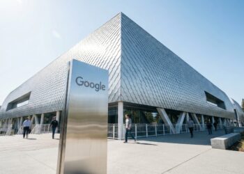 Una fotografía de ángulo amplio captura el distintivo techo de "piel solar" con escamas de dragón del edificio del campus Bay View de Google en Mountain View, California. Un gran letrero de metal cepillado con el logotipo de Google se encuentra en primer plano. Varias personas caminan por los senderos exteriores de la moderna estructura de vidrio y metal bajo un cielo despejado y soleado.