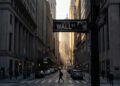 Fotografía urbana del letrero de la calle "WALL ST" en primer plano, en la intersección con Broad Street. La vista se adentra en el "cañón" de rascacielos del distrito financiero de Nueva York al atardecer. La luz dorada del sol se filtra entre los edificios al fondo, mientras la calle permanece en penumbra con peatones y tráfico. El estilo es documental y realista.
