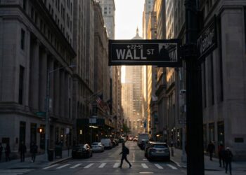 Fotografía urbana del letrero de la calle "WALL ST" en primer plano, en la intersección con Broad Street. La vista se adentra en el "cañón" de rascacielos del distrito financiero de Nueva York al atardecer. La luz dorada del sol se filtra entre los edificios al fondo, mientras la calle permanece en penumbra con peatones y tráfico. El estilo es documental y realista.