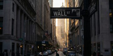 Fotografía urbana del letrero de la calle "WALL ST" en primer plano, en la intersección con Broad Street. La vista se adentra en el "cañón" de rascacielos del distrito financiero de Nueva York al atardecer. La luz dorada del sol se filtra entre los edificios al fondo, mientras la calle permanece en penumbra con peatones y tráfico. El estilo es documental y realista.