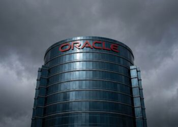 Fotografía de arquitectura en contrapicado de la sede corporativa de Oracle. El imponente edificio cilíndrico de cristal azulado se alza bajo un cielo denso y tormentoso de color gris plomo que se refleja en su fachada. El logotipo rojo corporativo de 'ORACLE' destaca en la parte superior de la estructura.