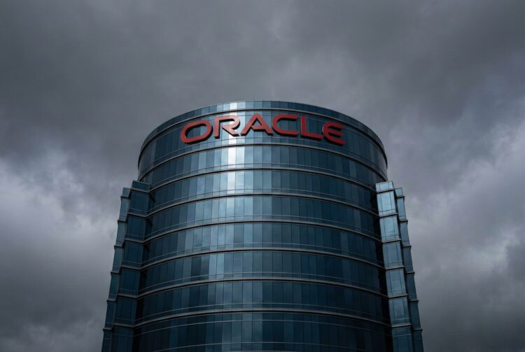 Fotografía de arquitectura en contrapicado de la sede corporativa de Oracle. El imponente edificio cilíndrico de cristal azulado se alza bajo un cielo denso y tormentoso de color gris plomo que se refleja en su fachada. El logotipo rojo corporativo de 'ORACLE' destaca en la parte superior de la estructura.