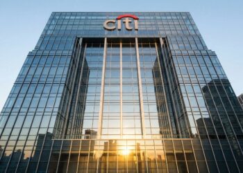 Fotografía de arquitectura en contrapicado de la fachada de cristal y acero de la sede mundial de Citigroup, ubicada en 388 Greenwich Street, Nueva York. La luz dorada del amanecer se refleja intensamente en los paneles inferiores del rascacielos, mientras que el logotipo rojo y azul de "citi" es claramente visible en la parte superior de la estructura bajo un cielo azul claro.