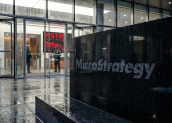 Fotografía exterior de la entrada del edificio de oficinas de MicroStrategy. En primer plano, un gran bloque de granito oscuro con el logotipo "MicroStrategy" grabado. El suelo está mojado por la lluvia, reflejando las luces del edificio y el entorno. Al fondo, se ve la entrada de cristal con dos guardias de seguridad en el vestíbulo.