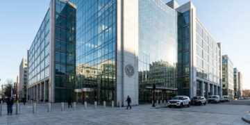 Fotografía panorámica exterior del edificio sede de la Comisión de Bolsa y Valores de EE. UU. (SEC) en Washington D.C. La moderna fachada de vidrio y piedra refleja la luz nítida de la mañana. El sello oficial de la SEC es visible en la pared de granito cerca de la entrada, con un vehículo policial estacionado y pocos peatones en la plaza adyacente.