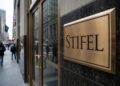 Una placa de bronce pulido con el logotipo de "STIFEL" grabado está montada en una pared de mármol oscuro en la entrada de un edificio de oficinas. Al fondo, se ve una acera urbana con peatones y edificios de la ciudad bajo un día nublado.