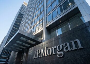Fotografía de arquitectura en contrapicado que muestra la imponente fachada de vidrio y acero de la sede mundial de JPMorgan Chase en Nueva York. El logotipo de "J.P. Morgan" en letras metálicas plateadas destaca sobre una base de granito oscuro en la entrada principal, bajo la luz de una mañana despejada.