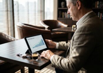 Fotografía de estilo de vida en un salón de negocios de lujo, donde un hombre de mediana edad con saco está sentado frente a una mesa de madera. Observa una tableta que muestra un gráfico de barras ascendente titulado "Recurring Revenue" (Ingresos Recurrentes). La luz natural se filtra a través de persianas venecianas, creando patrones de sombra. Un café espresso pequeño y un reloj de pulsera descansan sobre la mesa en primer plano. El fondo incluye sillones de cuero y estanterías borrosas.