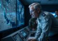 Un hombre de pelo gris con uniforme militar y gafas se inclina sobre una consola en un centro de mando con poca luz, mirando intensamente una gran pantalla que muestra un mapa estratégico del Golfo Pérsico, Irak y las regiones circundantes con varios iconos y datos. Otros miembros del personal militar están en el fondo. La iluminación proviene principalmente de las pantallas azules.
