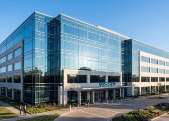 Una fotografía arquitectónica panorámica y profesional de la sede corporativa de Vistra Corp en Irving, Texas, en una mañana despejada. El edificio moderno de varios pisos combina una fachada de vidrio reflectante que muestra un cielo azul claro con detalles de piedra clara. El logotipo "VISTRA" está montado de forma elegante sobre la marquesina de la entrada principal. En primer plano, dos profesionales de negocios caminan por una plaza pavimentada rodeada de jardines bien cuidados y árboles frondosos. La luz del sol brillante ilumina la escena.