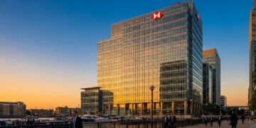 Fotografía exterior del rascacielos de la sede de HSBC (8 Canada Square) en los Docklands de Londres durante la puesta de sol. La fachada de vidrio del edificio refleja intensamente la luz cálida y dorada del sol. Profesionales con vestimenta de negocios caminan por el muelle en el primer plano, junto al agua y barcos atracados.
