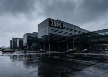 Fotografía arquitectónica panorámica del exterior de la sede corporativa de IBM en un día lluvioso. Un imponente edificio de cristal ahumado y acero muestra el clásico logotipo de IBM en su fachada superior derecha. El cielo gris plomo y el pavimento mojado, que refleja la estructura, crean una atmósfera fría, dramática y corporativa. Estilo editorial financiero.