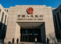 Fotografía exterior de la entrada principal de la sede del Banco Popular de China en Pekín. La fachada de piedra gris muestra el emblema nacional de China en rojo y dorado, y debajo el nombre "PEOPLE'S BANK OF CHINA" y "中国人民银行" en letras grandes. Varias personas con abrigos caminan frente al edificio bajo un cielo azul despejado.