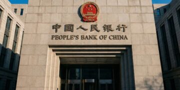 Fotografía exterior de la entrada principal de la sede del Banco Popular de China en Pekín. La fachada de piedra gris muestra el emblema nacional de China en rojo y dorado, y debajo el nombre "PEOPLE'S BANK OF CHINA" y "中国人民银行" en letras grandes. Varias personas con abrigos caminan frente al edificio bajo un cielo azul despejado.