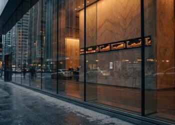 Vista desde el interior de un vestíbulo de oficina financiero moderno con paredes de mármol, mirando hacia una calle urbana mojada a través de grandes ventanas de cristal. Una pantalla LED montada en la pared muestra un gráfico de la cotización del "ORO" con una línea ascendente en tiempo real. Peatones borrosos y coches pasan por la acera y la calle bajo un cielo gris de la tarde, con rascacielos al fondo. La luz cálida del interior contrasta con el exterior húmedo.