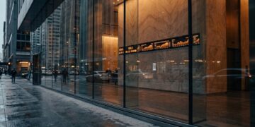 Vista desde el interior de un vestíbulo de oficina financiero moderno con paredes de mármol, mirando hacia una calle urbana mojada a través de grandes ventanas de cristal. Una pantalla LED montada en la pared muestra un gráfico de la cotización del "ORO" con una línea ascendente en tiempo real. Peatones borrosos y coches pasan por la acera y la calle bajo un cielo gris de la tarde, con rascacielos al fondo. La luz cálida del interior contrasta con el exterior húmedo.
