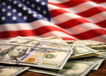 Billetes de dólar en primer plano con bandera de EE. UU. al fondo.