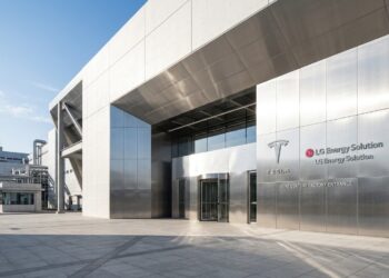Entrada de fábrica moderna con paneles metálicos y logotipos de Tesla y LG Energy Solution. Plaza pavimentada y cielo despejado.