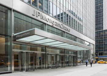 Entrada principal de vidrio y metal de un edificio de JPMorgan Chase con el nombre visible, personas caminando y un taxi amarillo al fondo.