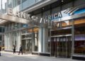 Un gran edificio moderno con fachada de vidrio y un letrero que dice "BANK OF AMERICA". Personas caminan por la acera de enfrente.