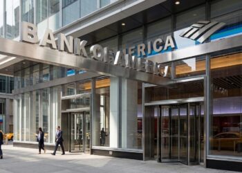 Un gran edificio moderno con fachada de vidrio y un letrero que dice "BANK OF AMERICA". Personas caminan por la acera de enfrente.