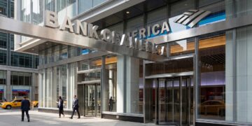 Un gran edificio moderno con fachada de vidrio y un letrero que dice "BANK OF AMERICA". Personas caminan por la acera de enfrente.